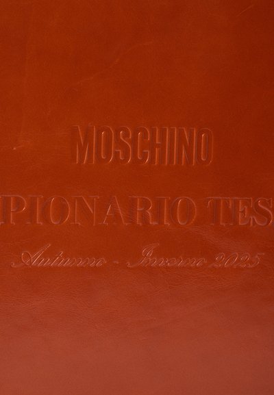 Surface en cuir marron avec du texte en relief : "MOSCHINO," "SPIANARIO TEST," et "Autunno - Inverno 2025." Texture lisse, design minimaliste.