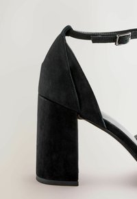 Černé semišové sandály na vysokém podpatku s robustním podpatkem a nastavitelným páskem kolem kotníku. Mají špičatou špičku a hladký, elegantní design.