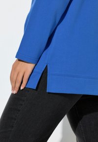 Modrý top s dlouhým rukávem a kulatým výstřihem, s bočními rozparky na spodním lemu a hladkou texturou. Spárovaný s černým denimem.