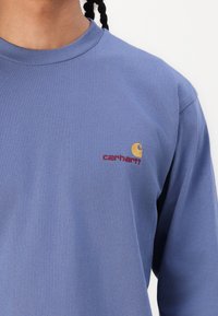 T-shirt à manches longues bleu en coton avec un col côtelé. Présente un petit logo brodé en jaune et rouge sur la poitrine.