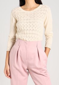 Jasno beżowy sweter z dzianiny z teksturowanym wzorem, okrągłym dekoltem i długimi rękawami, zestawiony z różowymi spodniami o wysokim stanie z kantami.