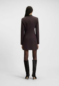 Vestito blazer corto viola con colletto nero, design aderente, maniche lunghe e texture liscia, abbinato a stivali neri alti fino al ginocchio.