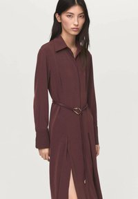 Vestito lungo bordeaux con maniche lunghe e colletto, dotato di uno spacco frontale, caratterizzato da una cintura in tessuto con fibbia dorata e una texture liscia e fluida.