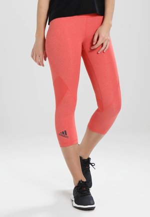 Vrouw draagt koraalkleurige Adidas cropped legging en zwarte sportschoenen, staand met één been gekruist voor het andere.