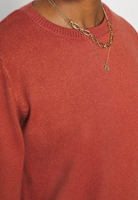 Maglione lavorato a maglia color ruggine con scollo rotondo, caratterizzato da una trama sottile. Accompagnato da collane in oro a strati.