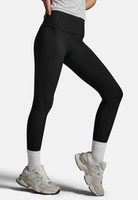 Czarne legginsy z wysokim stanem o gładkiej fakturze i bocznych szwach, w połączeniu z jasnymi butami sportowymi i białymi skarpetkami za kostkę.