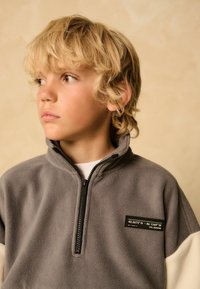 Grå fleece-pullover med høj krave, halv lynlås og kontrasterende cremede ærmer. Har et rektangulært sort mærke med koordinater.