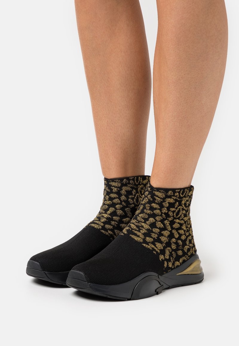 Jambes portant des baskets style chaussettes à imprimé léopard noir et or avec des semelles noires épaisses, debout sur fond blanc.