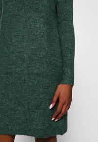 Grön stickad klänning med texturerad yta, långa ärmar och en lätt utsvängd fåll. Handen vilar på klänningen med manikyrerade naglar.