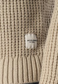 Primo piano di un tessuto a coste beige con un piccolo etichetta bianca che reca la scritta "JACK & JONES EST. 1990" cucita sulla superficie.