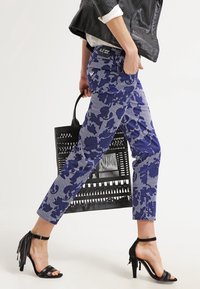 Marinblå byxor med blommönster, tillverkade av ett texturerat tyg, kombinerade med svarta sandaler med remmar och en perforerad svart tote-väska.