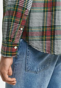Geruit shirt met groene, rode, gele en witte patronen, met knopen op de manchetten, gecombineerd met blauwe denim jeans.