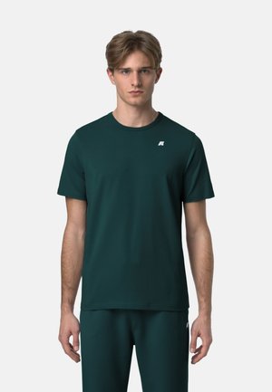 Giovane uomo con capelli castano chiaro che indossa una maglietta verde scuro a maniche corte e pantaloni abbinati, in piedi davanti a uno sfondo semplice.