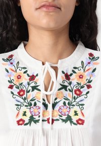 Blouse en coton blanc avec broderies florales en rouge, orange, violet et vert. Se noue au niveau du col, avec une texture froncée en bas.