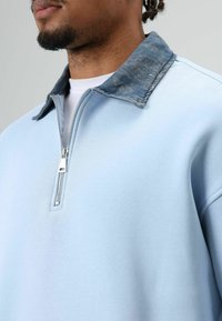 Sweatshirt bleu clair à demi-fermeture éclair avec col en denim. Tissu doux, coupe décontractée, texture contrastante du col et détail de fermeture éclair argentée.