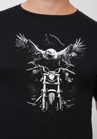 Černé tričko s grafickým potiskem motocyklu a orla kroužícího nad ním, ilustrované bílou barvou na tmavé látce.