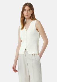 Crème mouwloos vest met een v-hals, voorzien van een knoopsluiting en een textuur gebreide stof. Gepaard met lichtbeige, losse broek.