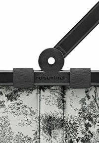 Poignée en métal noire attachée à un sac en textile à motif avec un design floral noir et blanc. Logo "reisenfolder" embossé sur la poignée.