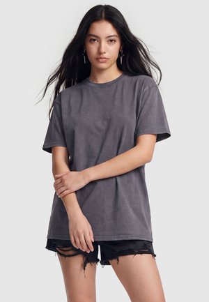 Ung kvinde med langt mørkt hår iført mørkegrå t-shirt, sorte revnede shorts og stjerneformede øreringe, står mod en lys baggrund.