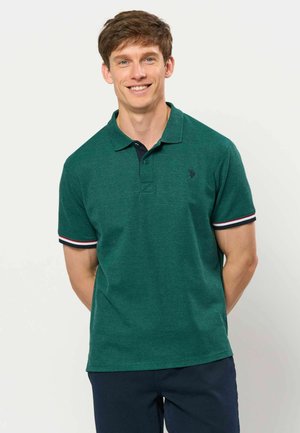 Jeune homme souriant portant un polo à manches courtes vert foncé avec des poignets rayés et un pantalon bleu foncé, debout les mains dans le dos.