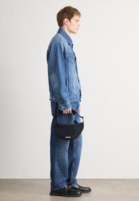 Jaqueta de denim com riscas verticais, combinada com jeans azuis largos. O modelo segura uma carteira preta com o logótipo "HUGO". Sapatos pretos completam o visual.