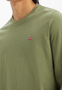 Grön bomullströja med långa ärmar och rundad halsringning, prydd med en röd Levi's logotyp på bröstet, med en slät textur och avslappnad passform.