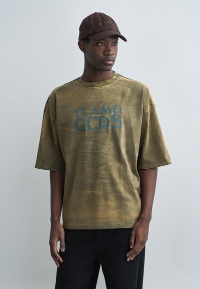 T-shirt oversize verde oliva con maniche corte, con la scritta "TI AMO GCDS" in grassetto blu. Pantaloni neri e un cappello marrone completano il look.
