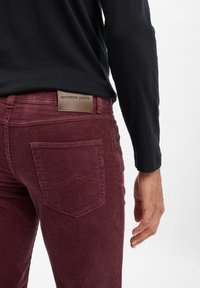 Mann in burgunderfarbenen Cordhosen mit Gesäßtasche und Leder-Label, schwarzem Langarmshirt, Hand entspannt an der Seite.
