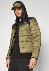 Giacca a vento verde oliva con dettagli neri, zip intera e cappuccio. Presenta un logo sul petto e cuciture orizzontali testurizzate.