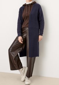 Dunkelblaue Strickjacke mit gerippter Textur, offener Front, getragen über einem braunen Hoodie und glänzenden braunen Weitbein-Hosen aus Kunstleder mit beigen Sneakers.