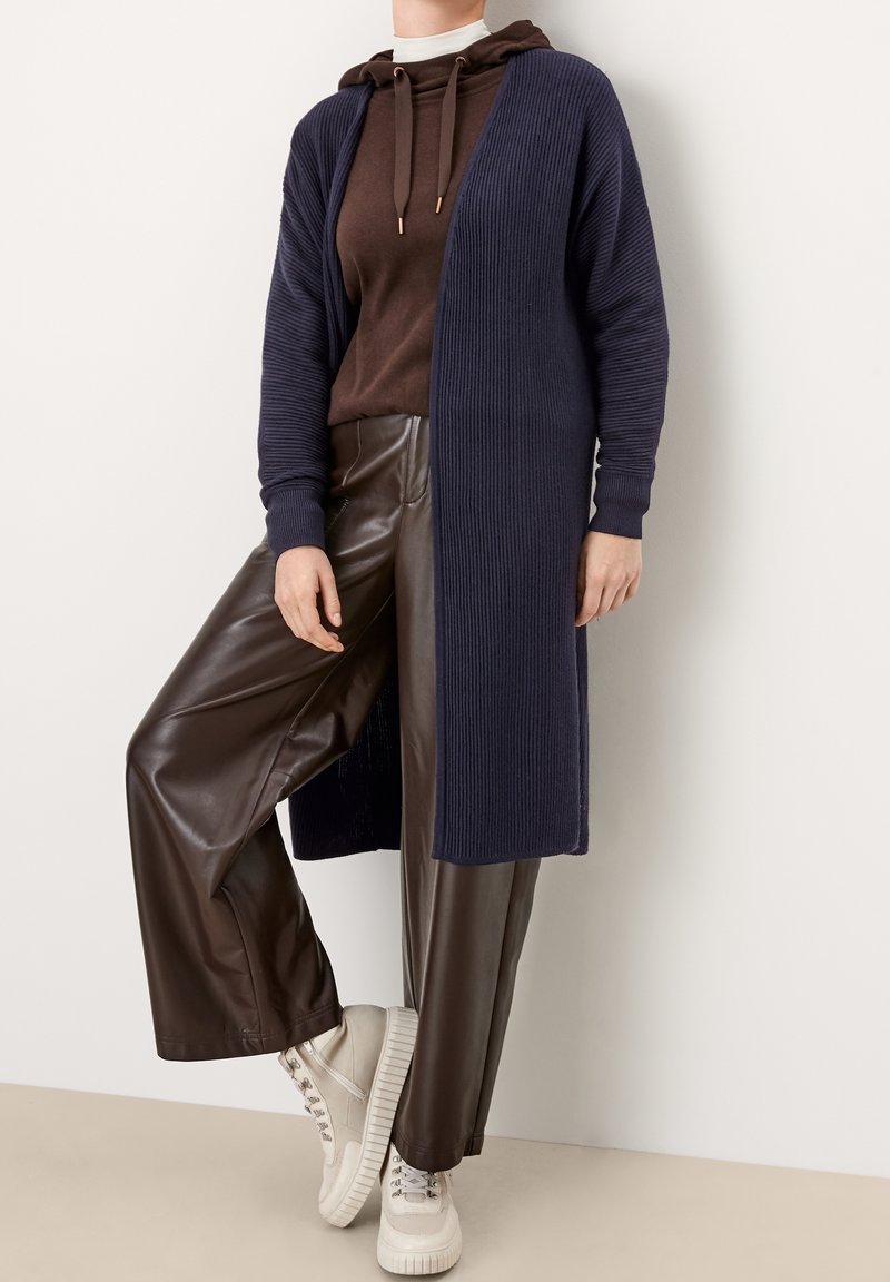 Dunkelblaue Strickjacke mit gerippter Textur, offener Front, getragen über einem braunen Hoodie und glänzenden braunen Weitbein-Hosen aus Kunstleder mit beigen Sneakers.