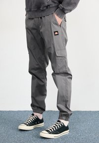 Pantalons cargo gris en tissu léger, avec poches latérales, des poignets élastiques et un petit logo cousu. Associés à des baskets noires.