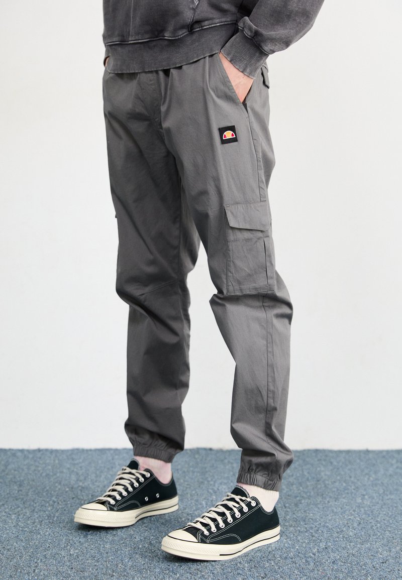 Pantalons cargo gris en tissu léger, avec poches latérales, des poignets élastiques et un petit logo cousu. Associés à des baskets noires.