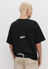 Sort oversized t-shirt lavet af bomuld, med et hvidt logo på ryggen. Korte ærmer og en afslappet pasform. Bæres med beige bukser.