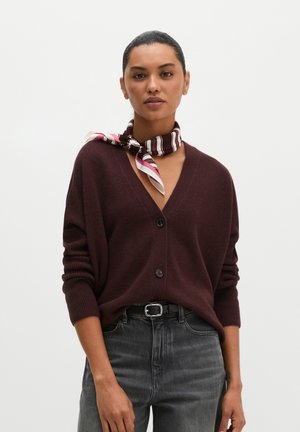 Frau mit zurückgekämmtem Haar trägt eine dunkelbraune, geknöpfte Strickjacke, schwarzen Gürtel, graue Jeans und einen gestreiften Seidenschal, der um ihren Hals gebunden ist.