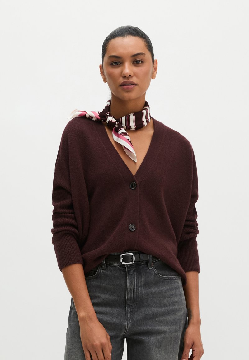 Woman with slicked-back hair wears a dark brown buttoned cardigan, black belt, gray jeans, and a striped silk scarf tied around her neck.
