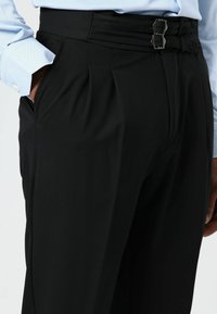 Pantalon noir ajusté avec plis, confectionné en tissu lisse. Doté d'une fermeture unique à double boucle et de poches latérales.