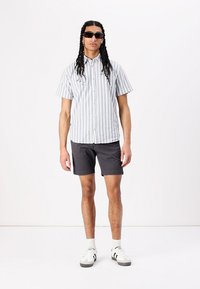 Short-sleeve striped shirt in white and blue, paired with gray shorts and black sunglasses. Model wears white sneakers with black accents.
