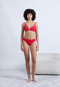 Rdeč čipkast modrček s cvetličnim vzorcem in usklajene rdeče hlačke, oba iz gladkega materiala. Model stoji bosa proti svetli ozadju.