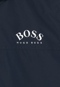 Tejido azul marino con un patrón texturizado. "BOSS" en negrita con una fuente serif blanca y "HUGO BOSS" en una fuente más pequeña debajo, centrado.