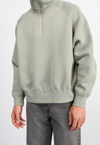 Ljusgrön sweatshirt med hög krage och kvart zip. Tillverkad av mjukt tyg, har ribbade ärmslut och fåll, tillsammans med grå jeans.