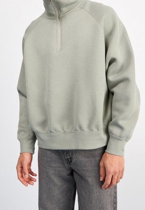 Felpa verde chiaro con colletto alto e zip a quarto. Realizzata in un tessuto morbido, presenta polsini e orlo a costine, abbinata a jeans grigi.