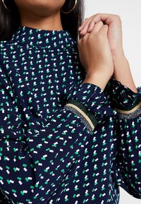 Blouse bleu marine avec un motif géométrique vert et blanc, dotée d'un col montant, de manches évasées et d'accents texturés aux poignets en or et argent.