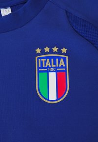 Plava dukserica s logotipom u obliku štita, ukrašenim zlatnim detaljima i četiri zlatne zvjezdice, s natpisom "ITALIA" i bojama talijanske zastave.