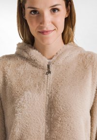 Sudadera beige de felpa con cremallera, de textura suave, que cuenta con capucha y cierre de cremallera, diseñada para un ajuste cómodo.