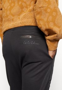 Sorte bukser med en baglomme, der har en tryklås og broderet "Carla Colucci" logo. Materialet ser glat ud med en let glans.