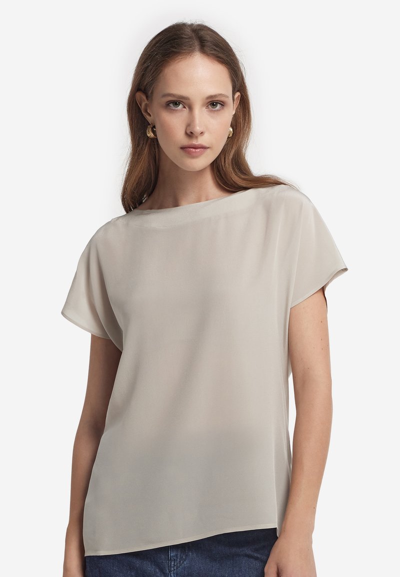 Jeune femme aux cheveux châtain clair portant des boucles d'oreilles créoles en or et un haut beige ample à manches courtes, debout devant un fond uni.