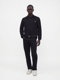 Homme debout portant un polo noir à manches longues, un pantalon noir avec ceinture et des baskets blanches sur fond blanc uni.