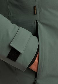 Giacca verde in tessuto testurizzato, con tasca con zip e regolazione al polsino in Velcro. Etichetta del marchio visibile vicino alla cucitura della spalla.