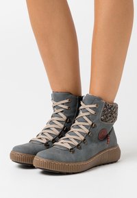 Botas altas de ante gris con cordones beige, cuello de punto texturizado y suela de goma con un logo circular y detalle en rojo.